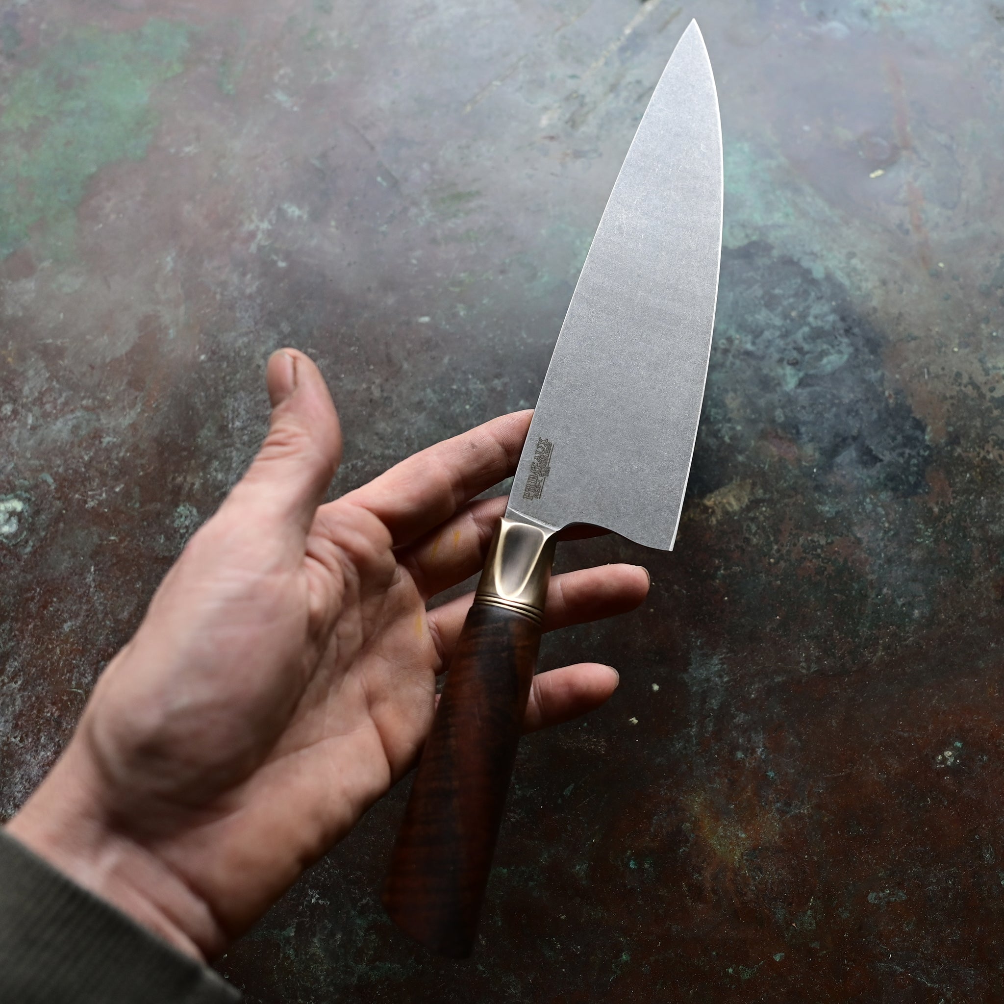 A chef knife with a stainless AEB-L steel blade, aged bronze bolster, and Claro Walnut handle, resting on a concrete surface