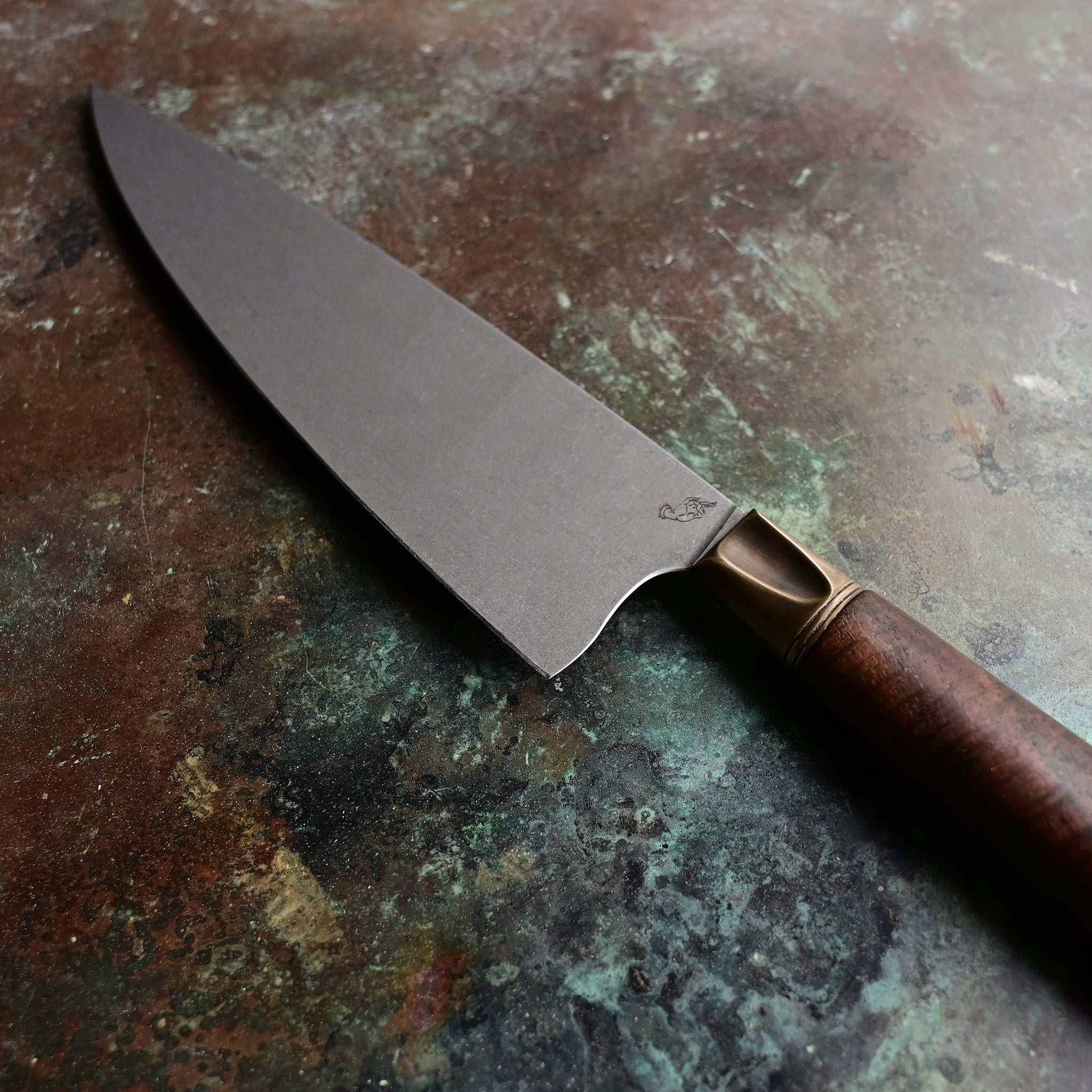 A chef knife with a stainless AEB-L steel blade, aged bronze bolster, and Claro Walnut handle, resting on a concrete surface