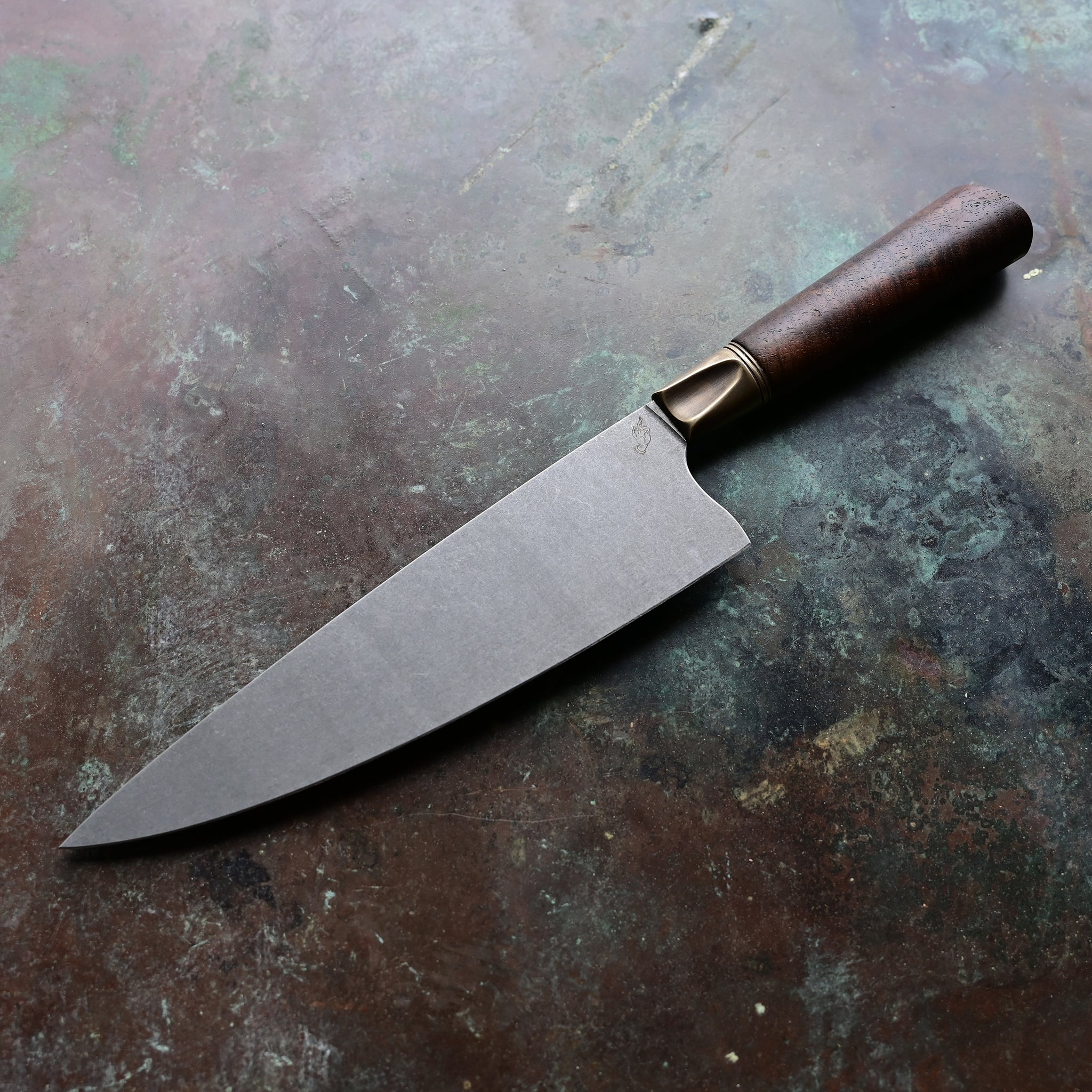 A chef knife with a stainless AEB-L steel blade, aged bronze bolster, and Claro Walnut handle, resting on a concrete surface