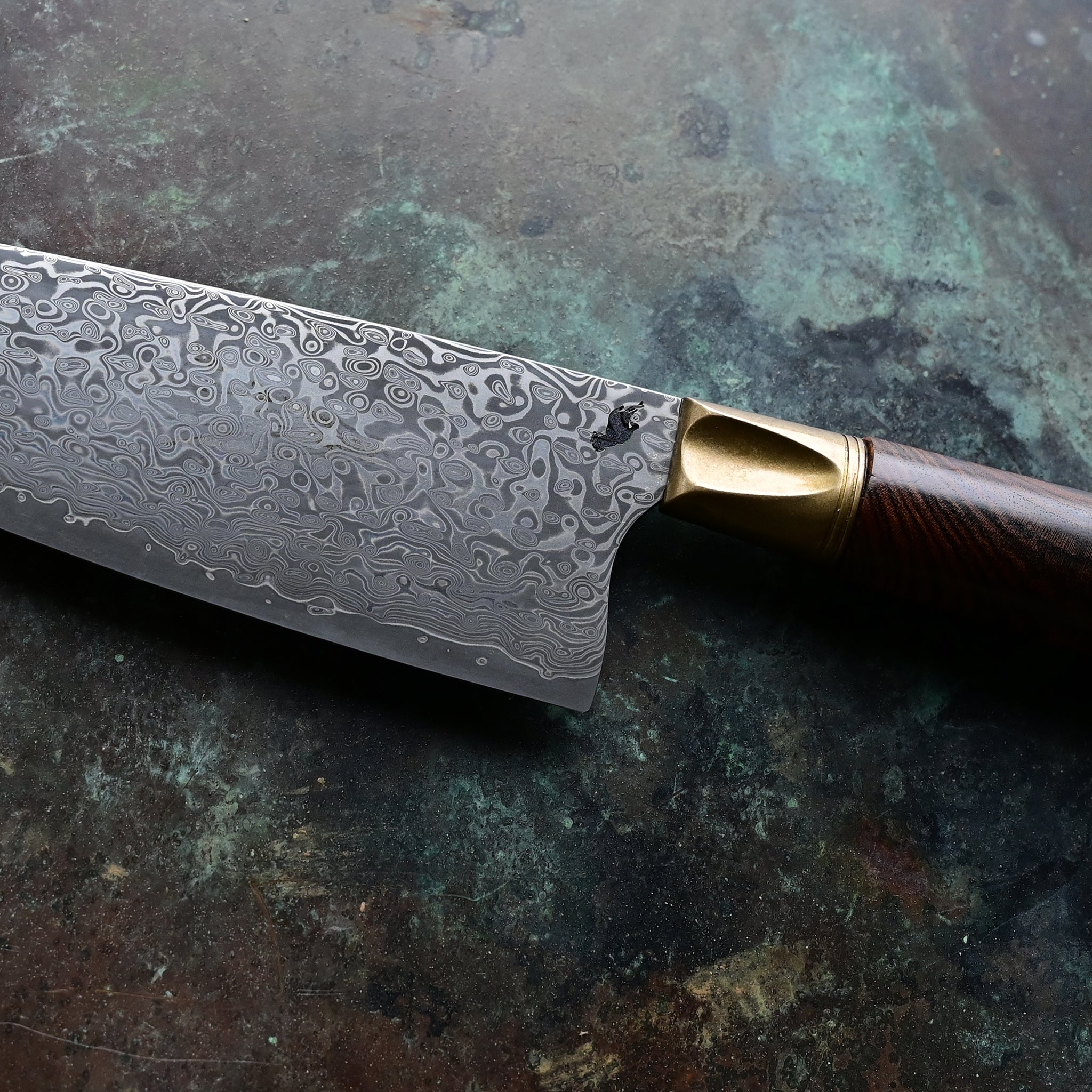 A Santoku knife with a starfall Damascus pattern, natural bronze bolster, and stabilized golden Buckeye burl handle, on a concrete backdrop