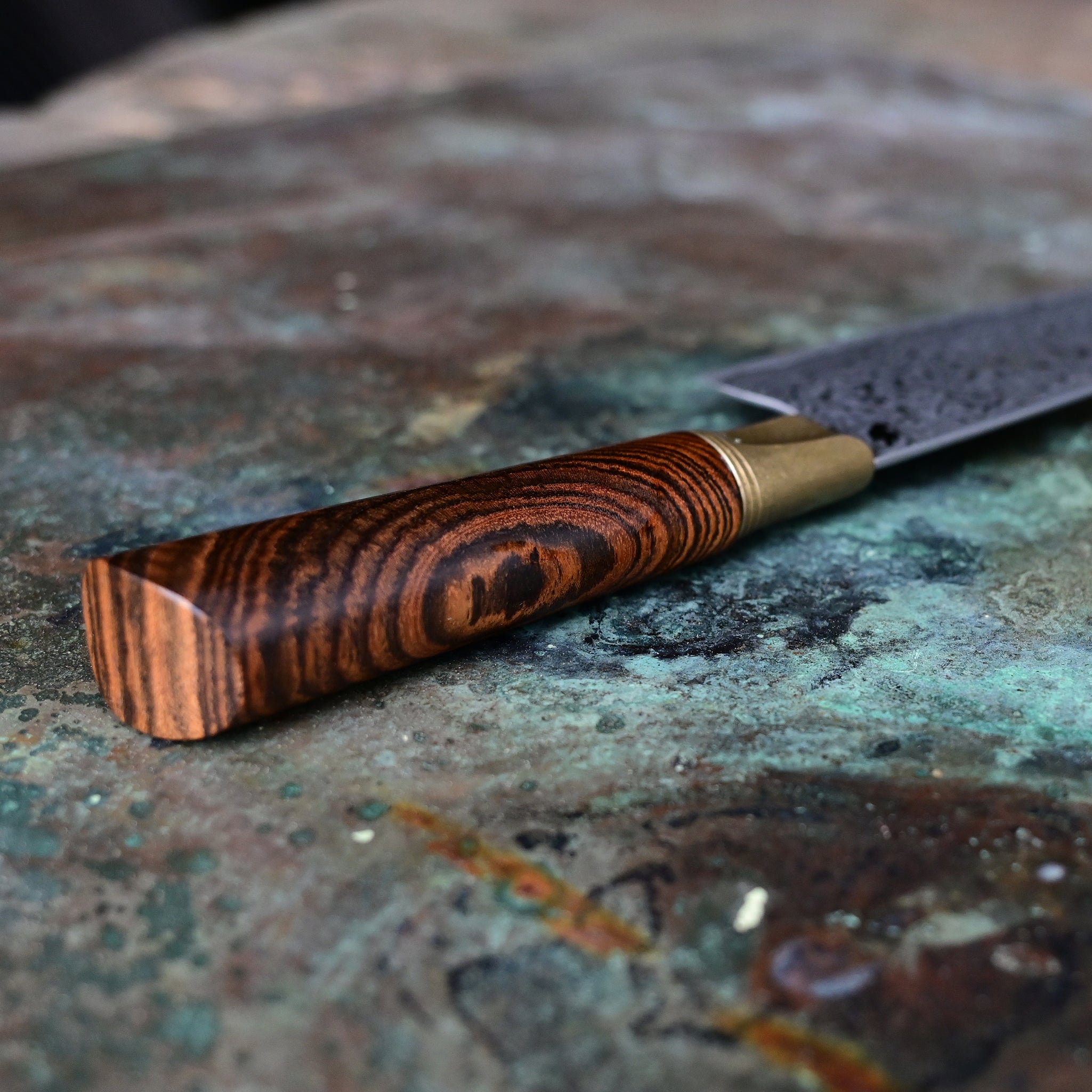 A Santoku knife with a starfall Damascus pattern, natural bronze bolster, and stabilized golden Buckeye burl handle, on a concrete backdrop