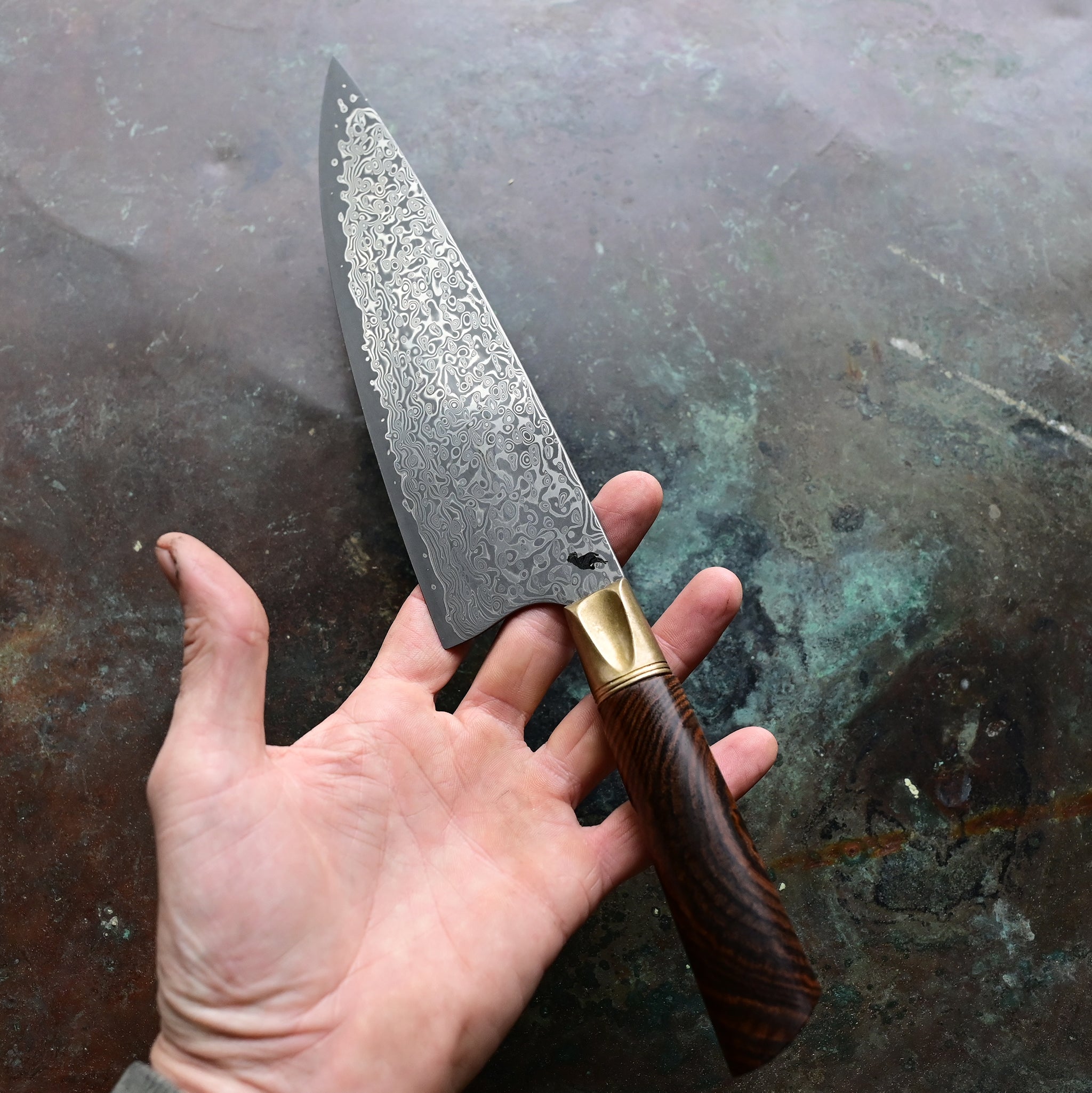A Santoku knife with a starfall Damascus pattern, natural bronze bolster, and stabilized golden Buckeye burl handle, on a concrete backdrop