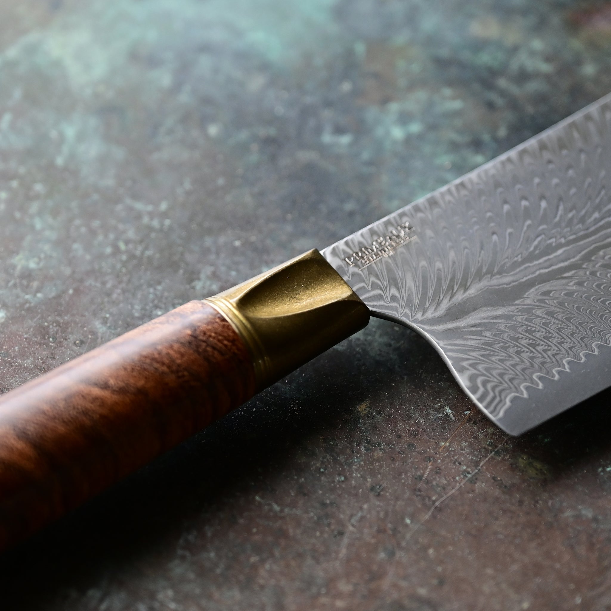 A mini cleaver with a bronze bolster, feather Damascus VG10 blade, and a Bocote wood handle, showcased on a concrete background