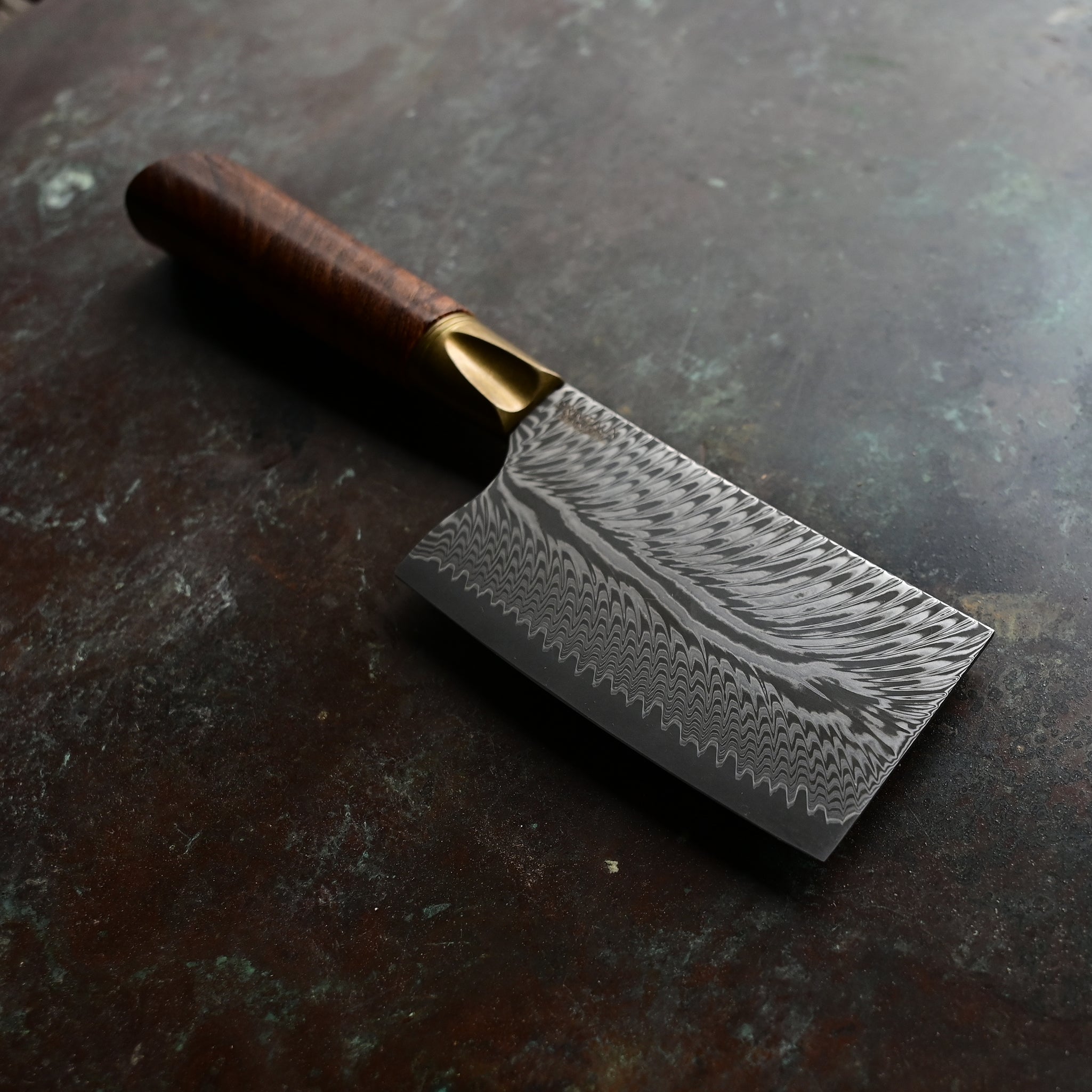 A mini cleaver with a bronze bolster, feather Damascus VG10 blade, and a Bocote wood handle, showcased on a concrete background
