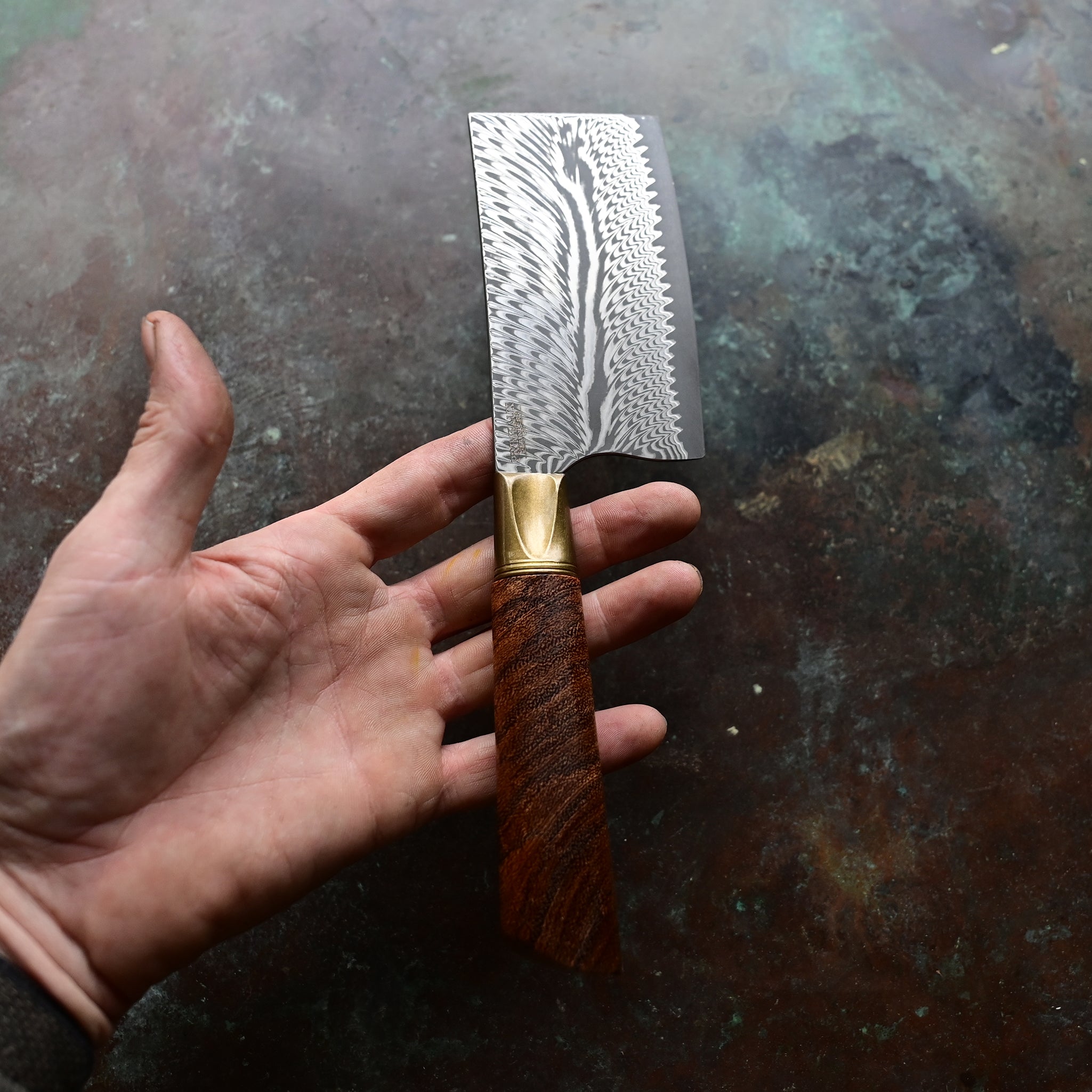 A mini cleaver with a bronze bolster, feather Damascus VG10 blade, and a Bocote wood handle, showcased on a concrete background