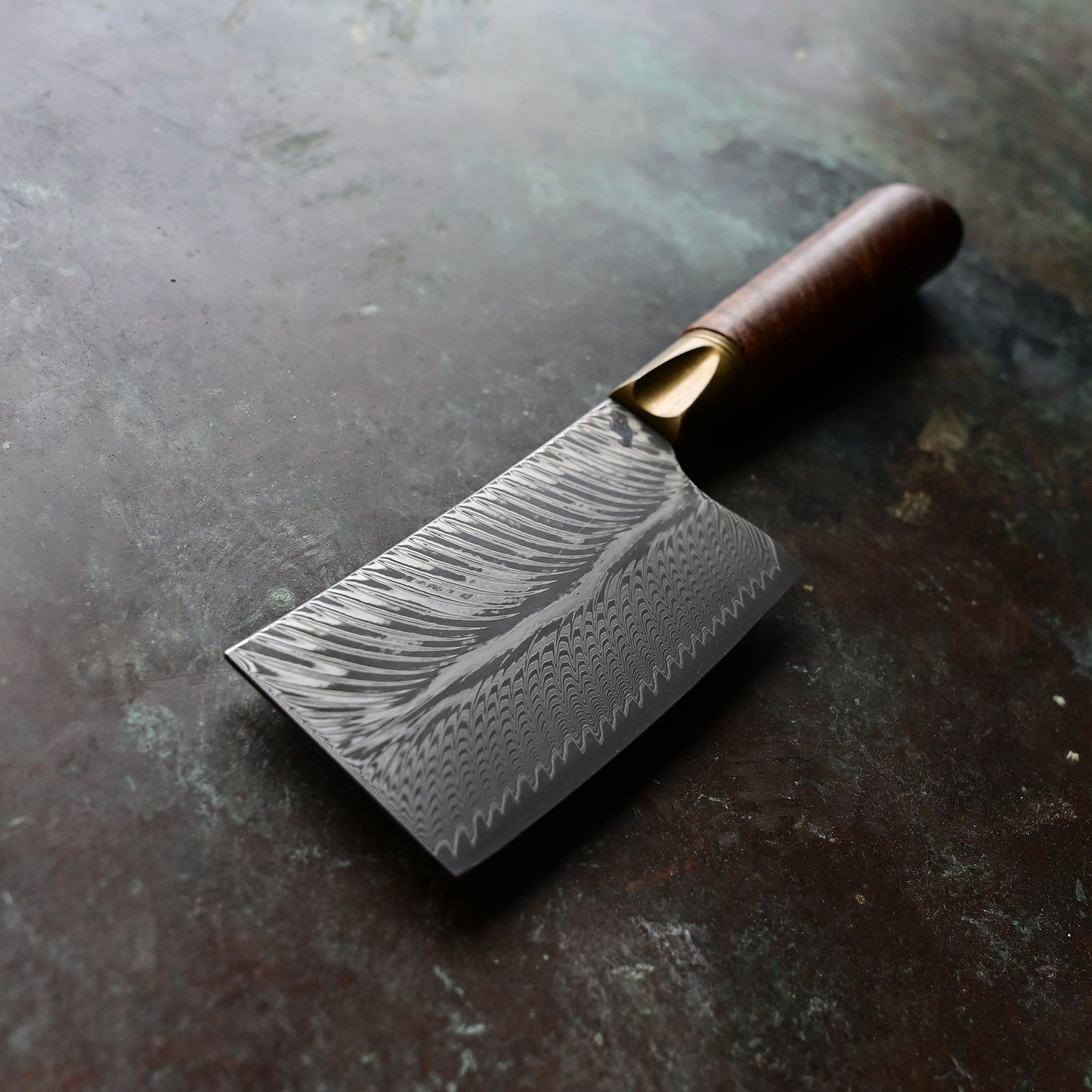 A mini cleaver with a bronze bolster, feather Damascus VG10 blade, and a Bocote wood handle, showcased on a concrete background