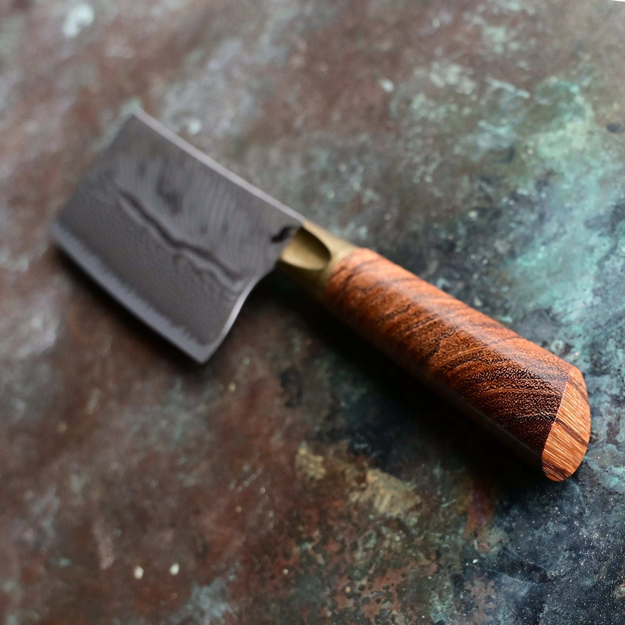 A mini cleaver with a bronze bolster, feather Damascus VG10 blade, and a Bocote wood handle, showcased on a concrete background
