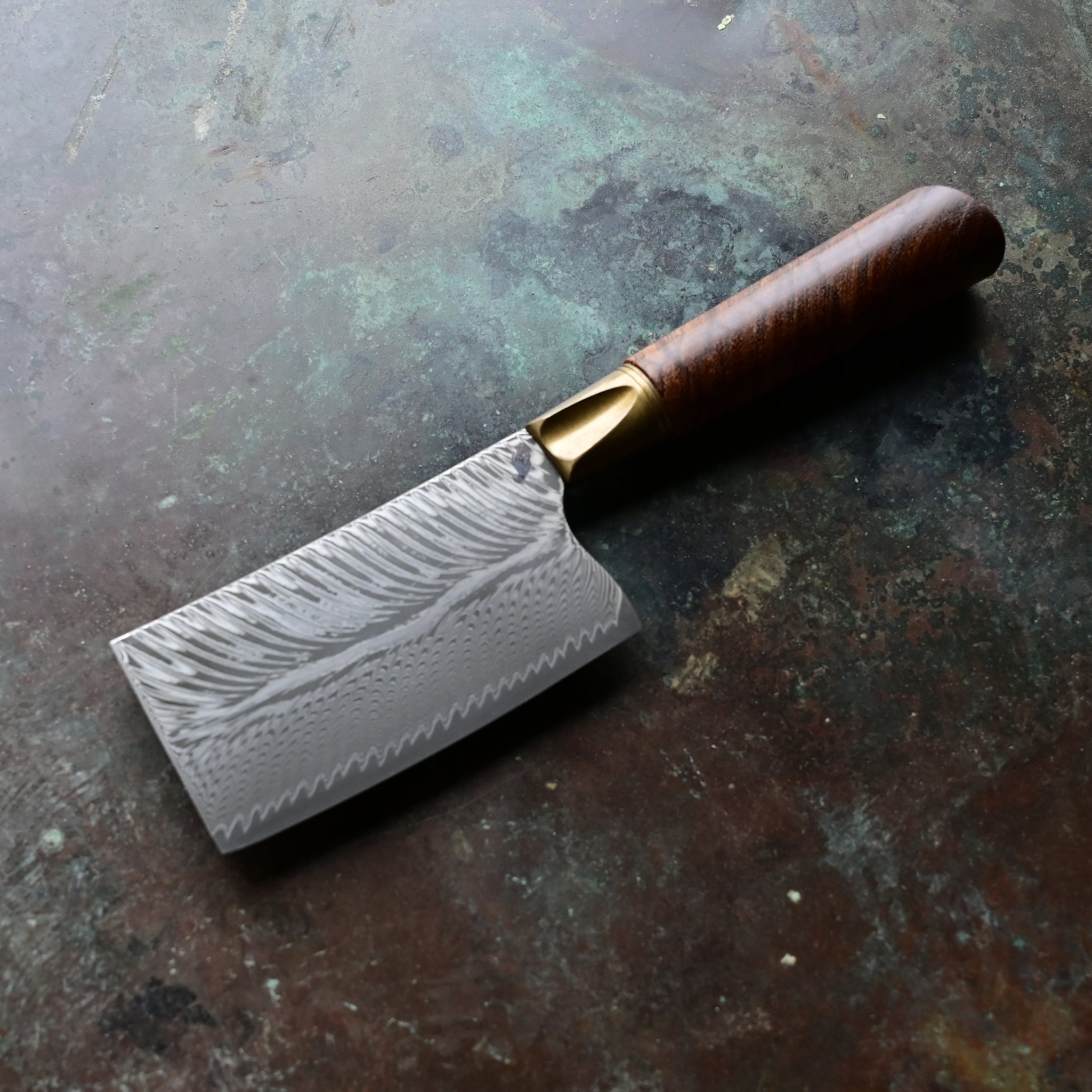 A mini cleaver with a bronze bolster, feather Damascus VG10 blade, and a Bocote wood handle, showcased on a concrete background