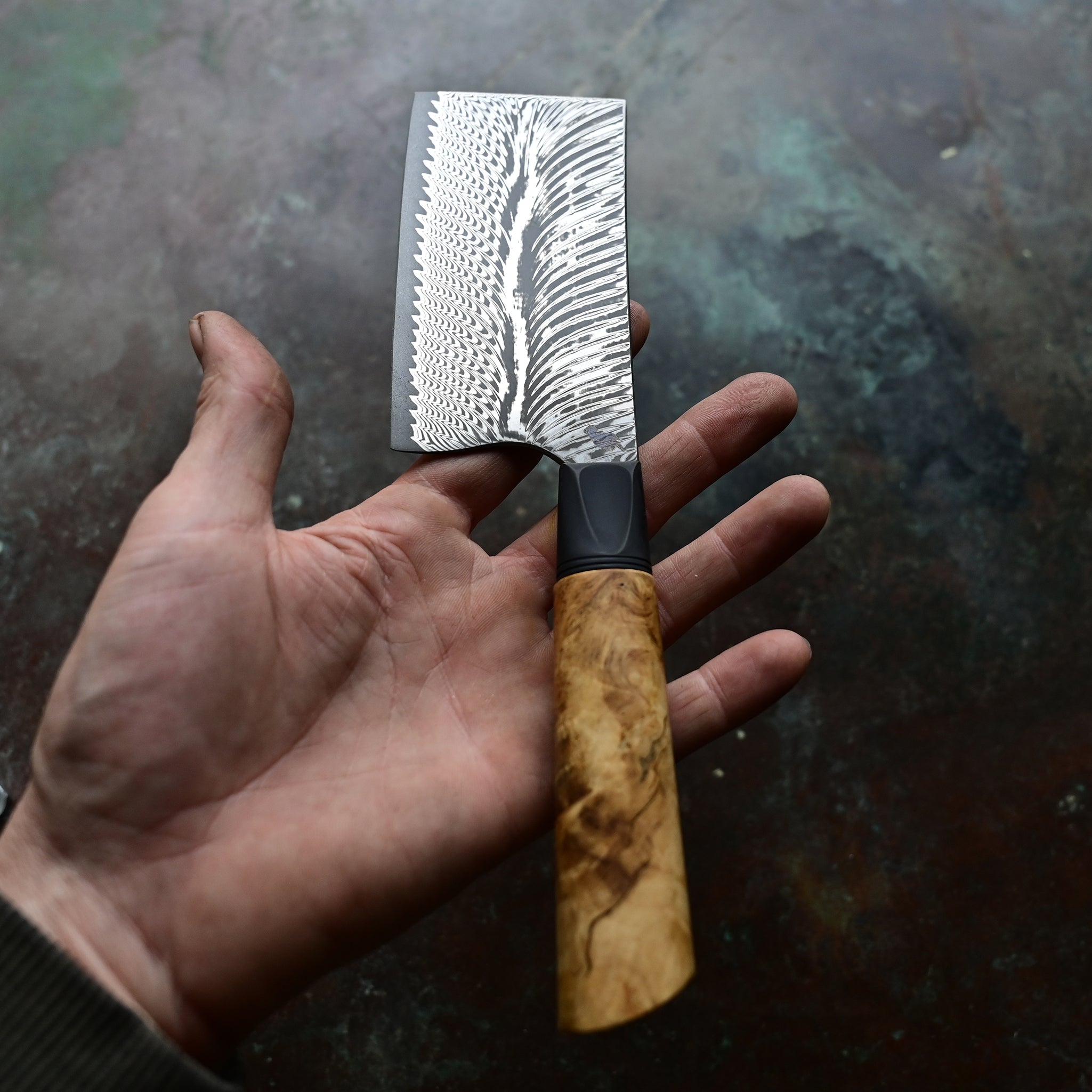 Unique mini cleaver on a concrete background, featuring a 4.5" long feather Damascus VG10 stainless blade, a sleek black anodized bolster, and a stabilized golden Buckeye burl wood handle with intricate patterns and color variations.
