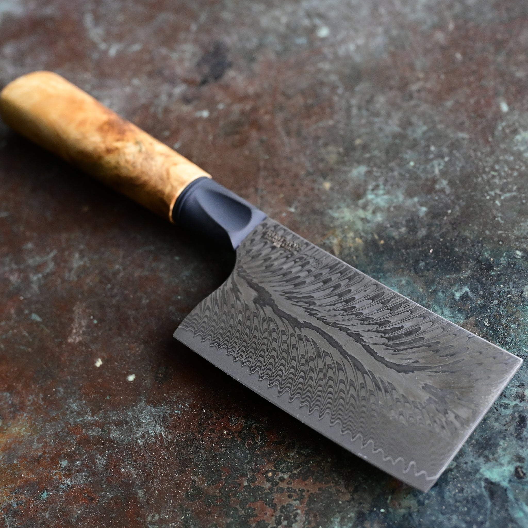 Unique mini cleaver on a concrete background, featuring a 4.5" long feather Damascus VG10 stainless blade, a sleek black anodized bolster, and a stabilized golden Buckeye burl wood handle with intricate patterns and color variations.