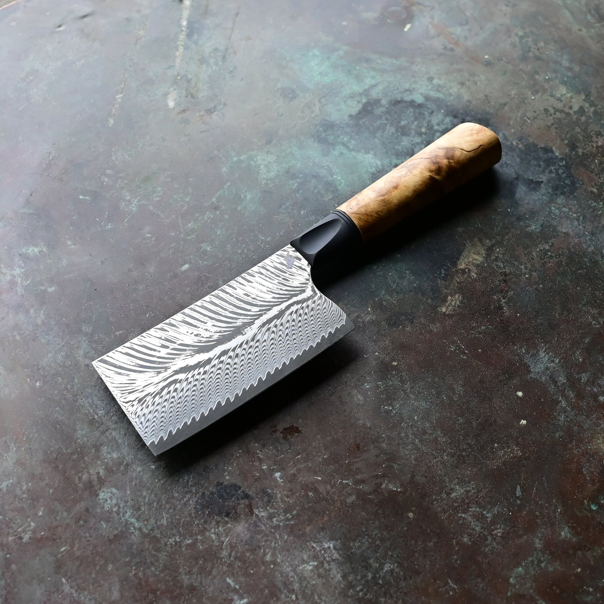 Unique mini cleaver on a concrete background, featuring a 4.5" long feather Damascus VG10 stainless blade, a sleek black anodized bolster, and a stabilized golden Buckeye burl wood handle with intricate patterns and color variations.