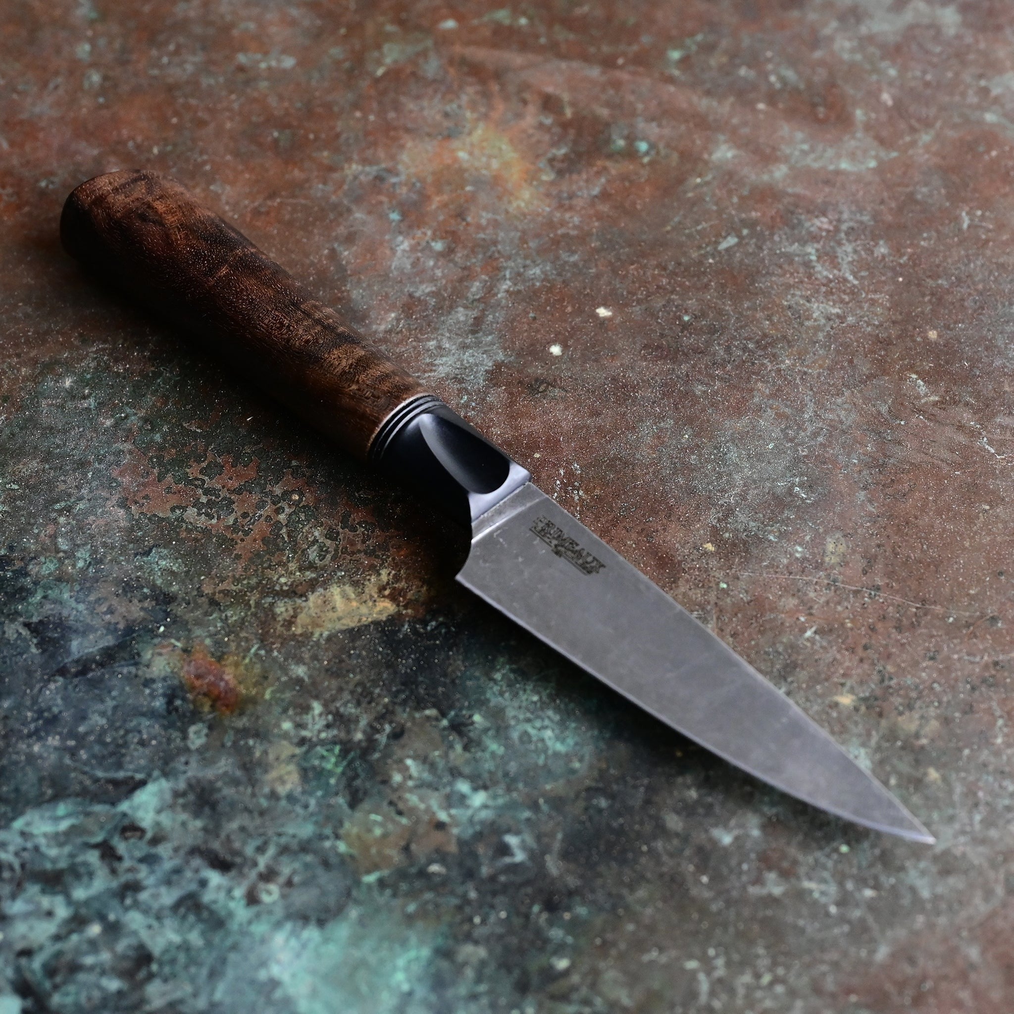 Paring knife with a deep, dark stone wash finish blade on a concrete backdrop, complemented by a black anodized bolster and a rich brown Claro walnut handle, featuring a thin, razor-sharp AEBL blade.