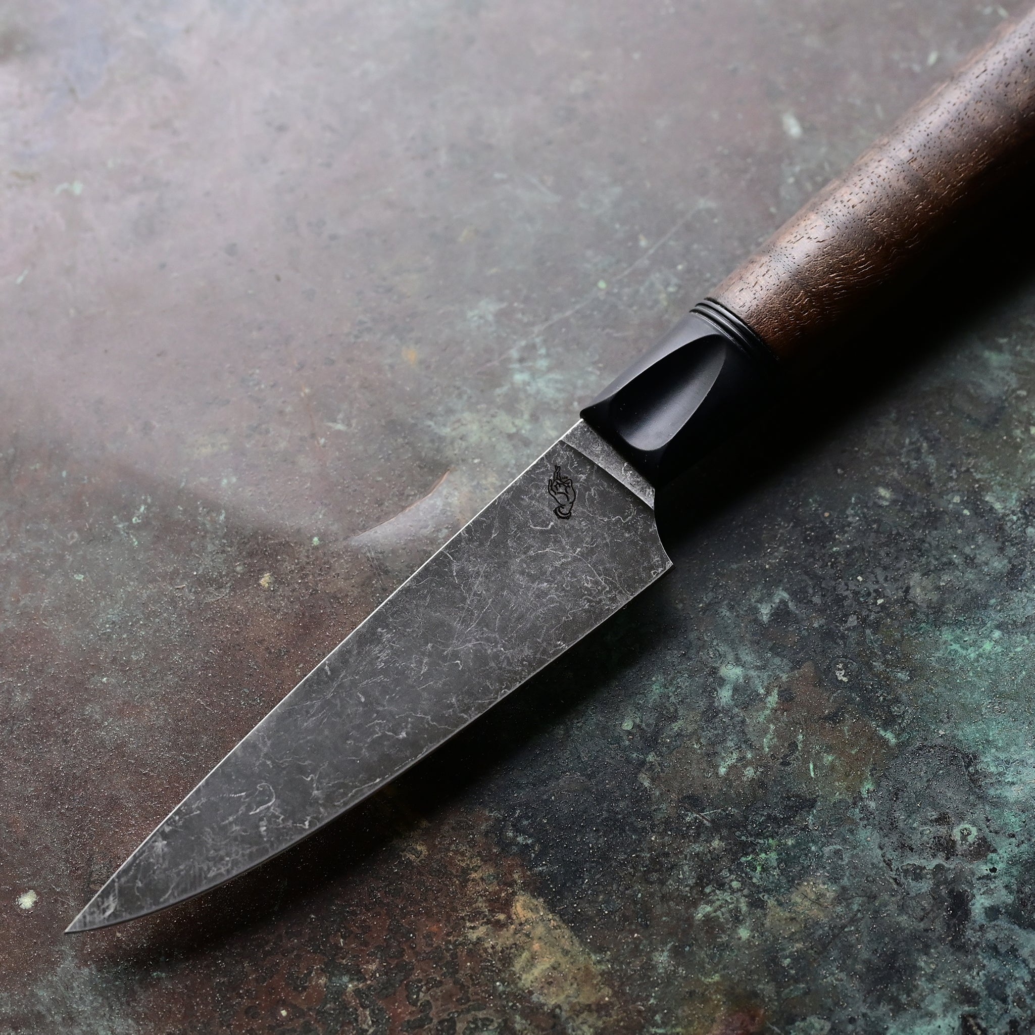 Paring knife with a deep, dark stone wash finish blade on a concrete backdrop, complemented by a black anodized bolster and a rich brown Claro walnut handle, featuring a thin, razor-sharp AEBL blade.