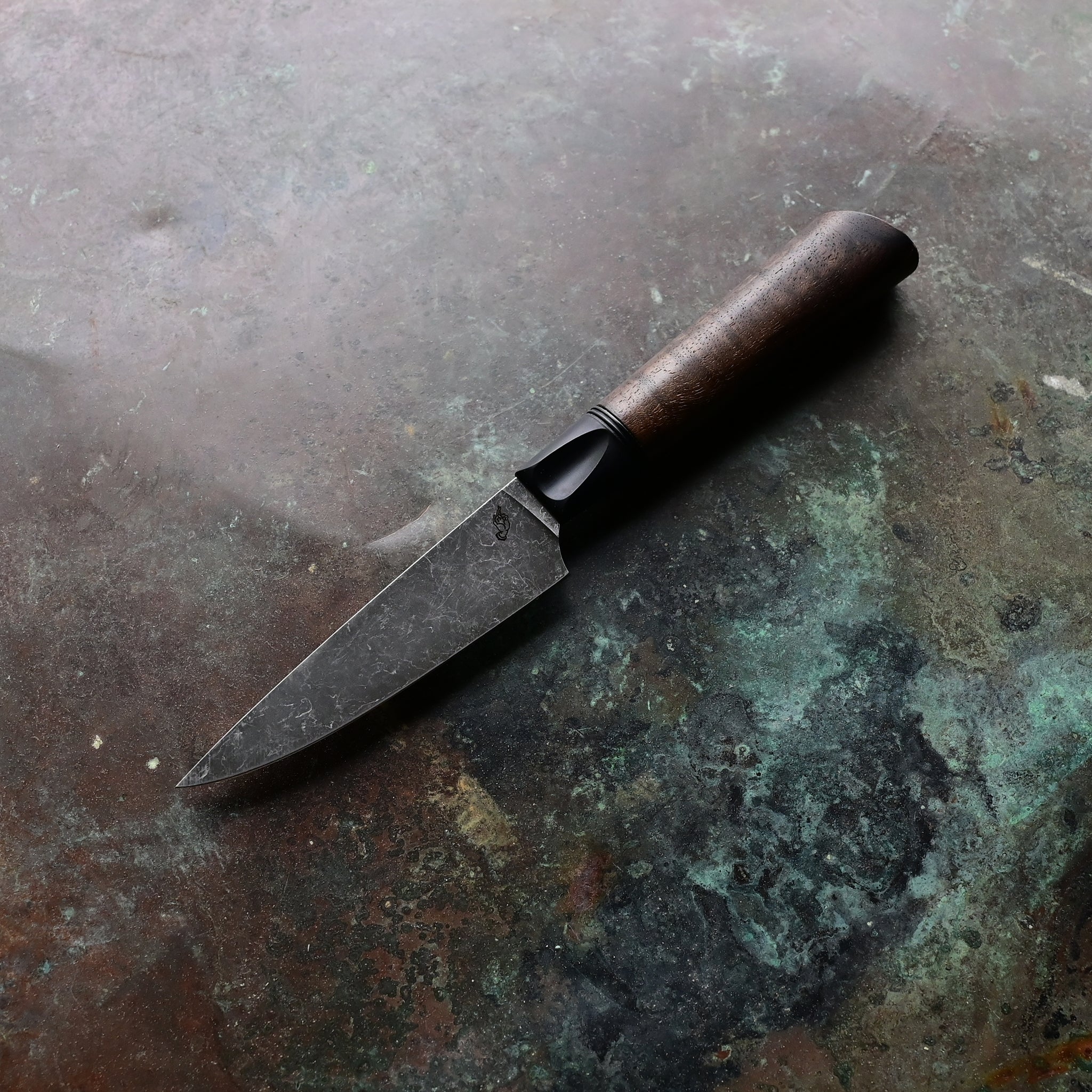 Paring knife with a deep, dark stone wash finish blade on a concrete backdrop, complemented by a black anodized bolster and a rich brown Claro walnut handle, featuring a thin, razor-sharp AEBL blade.