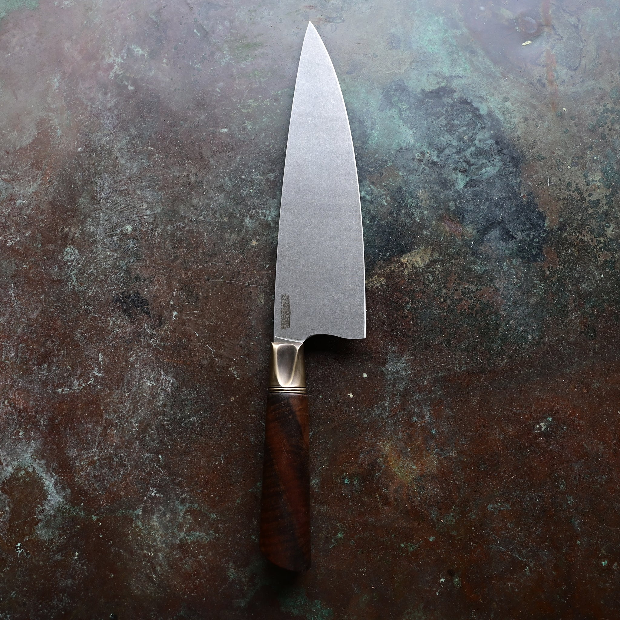 A chef knife with a stainless AEB-L steel blade, aged bronze bolster, and Claro Walnut handle, resting on a concrete surface