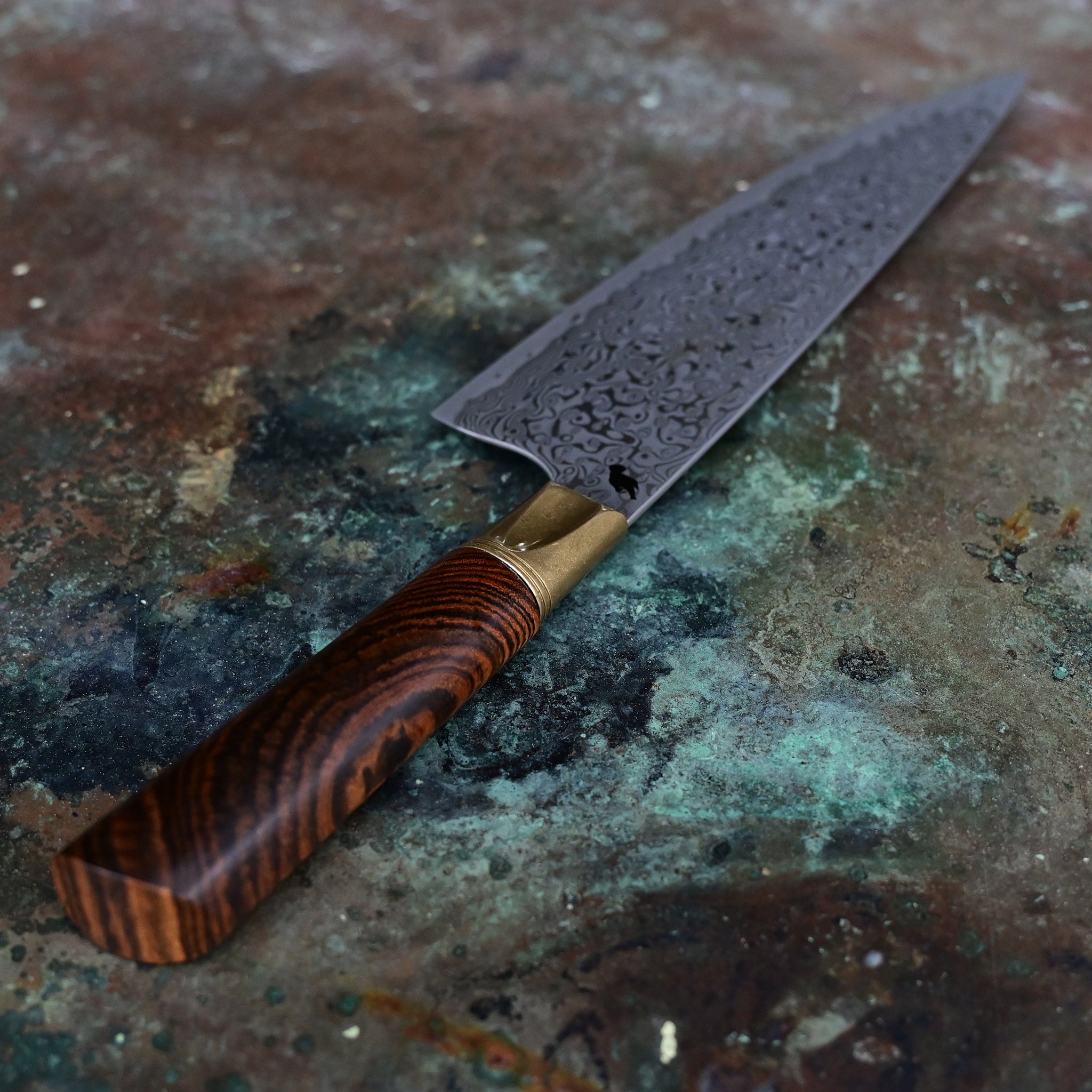 A stunning chef knife with a VG10 stainless Damascus zenith blade, aged bronze bolster, and a beautiful cross-cut Bocote wood handle displaying rings and patterns in shades of brown and tan, hand-forged by John Phillips