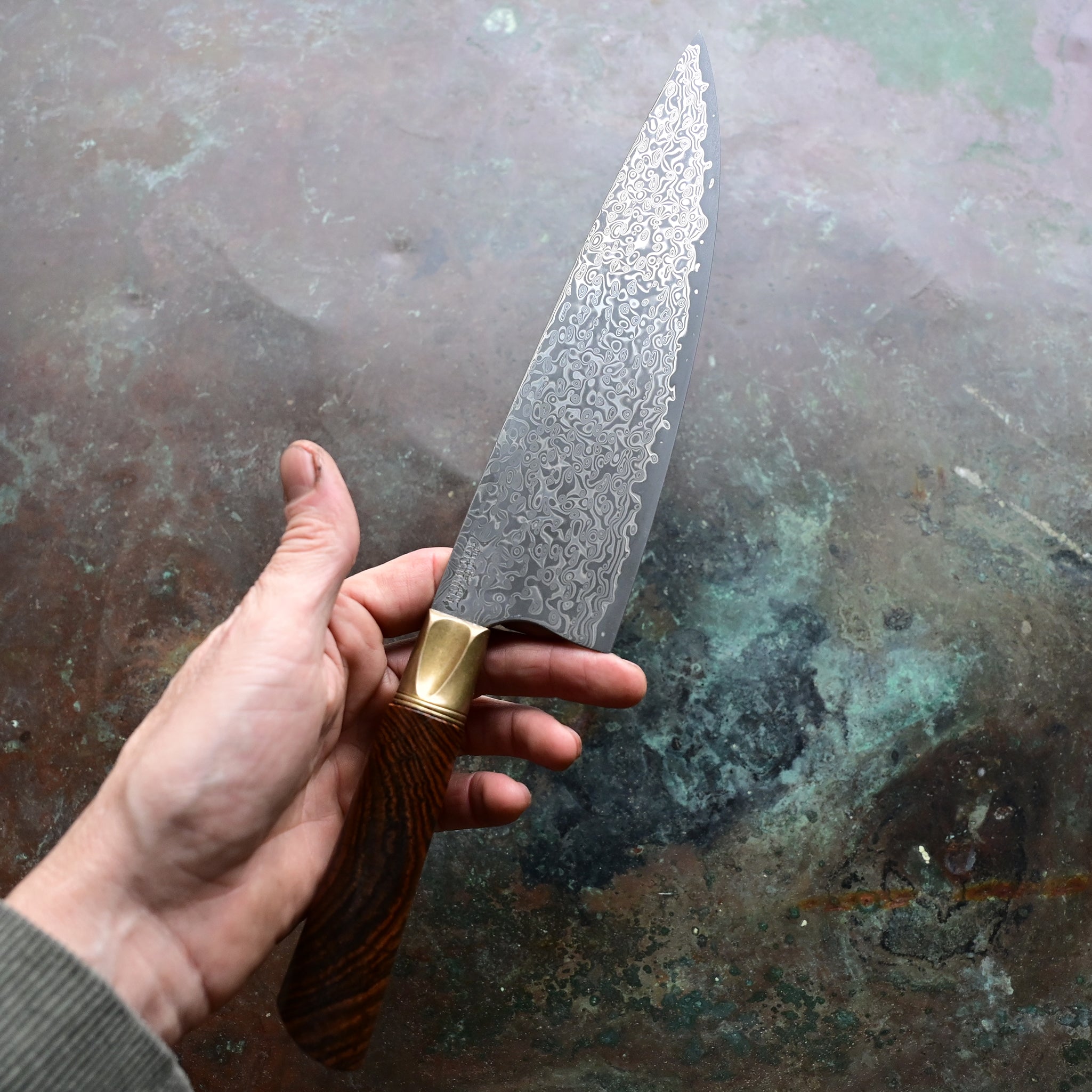 A Santoku knife with a starfall Damascus pattern, natural bronze bolster, and stabilized golden Buckeye burl handle, on a concrete backdrop