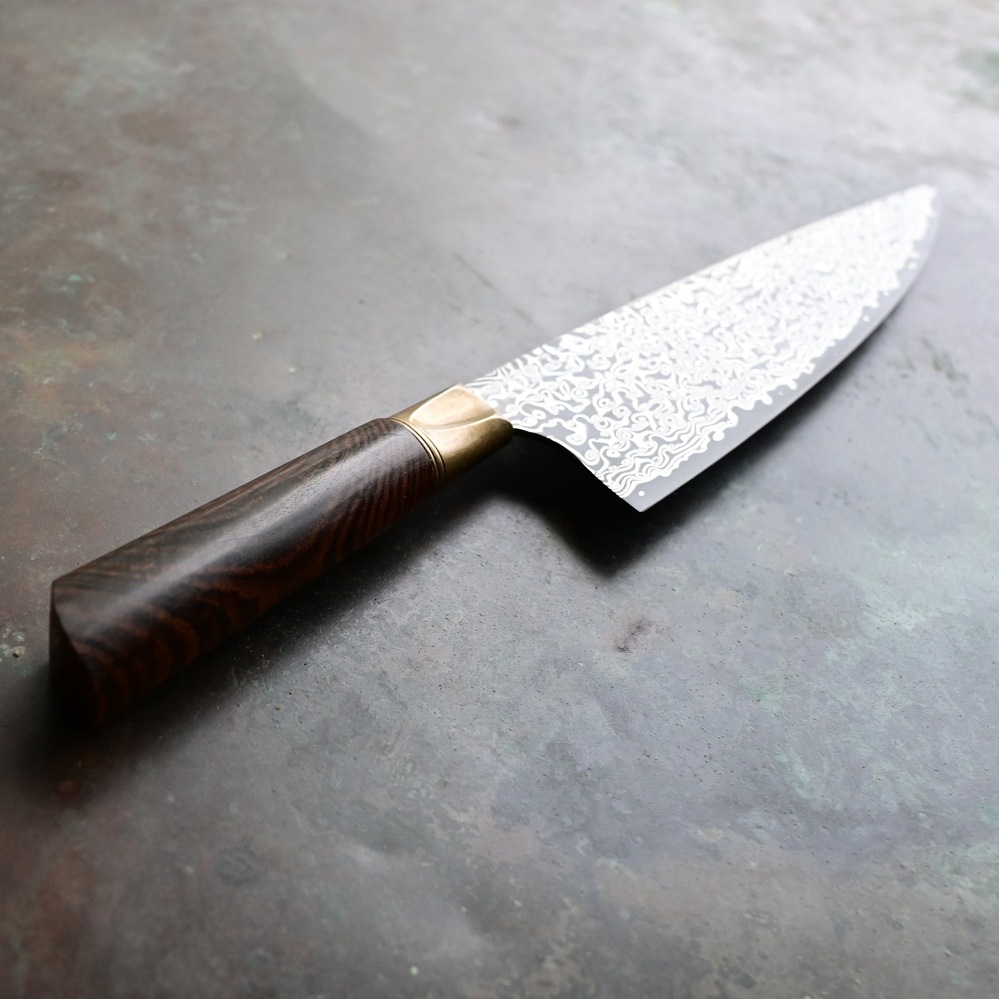 A Santoku knife with a starfall Damascus pattern, natural bronze bolster, and stabilized golden Buckeye burl handle, on a concrete backdrop