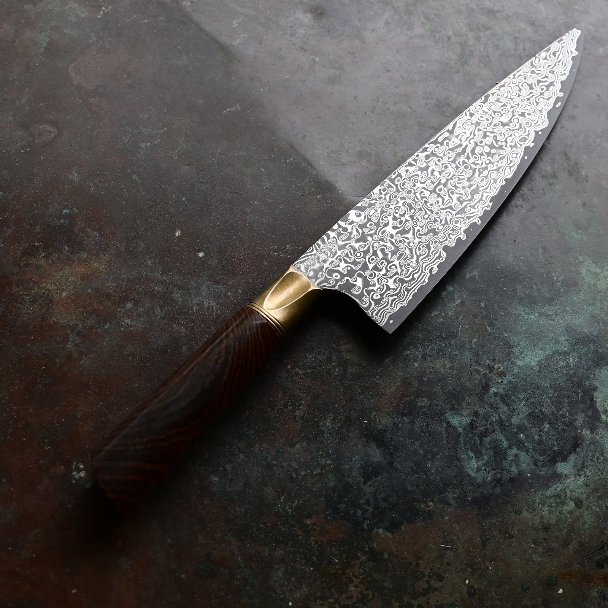 A stunning chef knife with a VG10 stainless Damascus zenith blade, aged bronze bolster, and a beautiful cross-cut Bocote wood handle displaying rings and patterns in shades of brown and tan, hand-forged by John Phillips