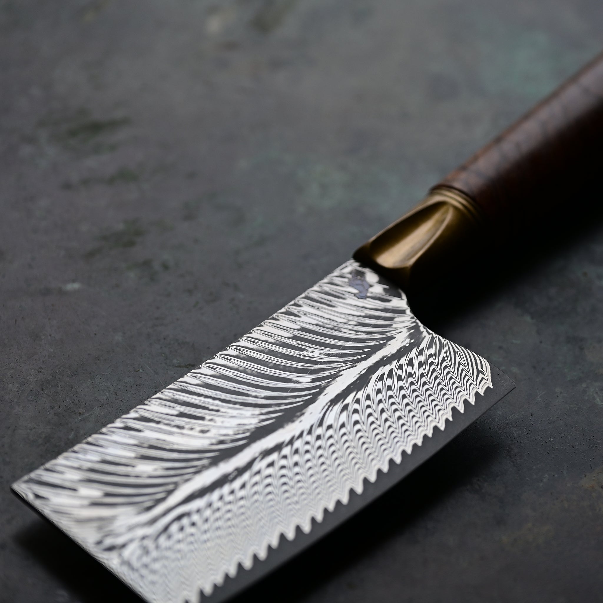 A mini cleaver with a bronze bolster, feather Damascus VG10 blade, and a Bocote wood handle, showcased on a concrete background