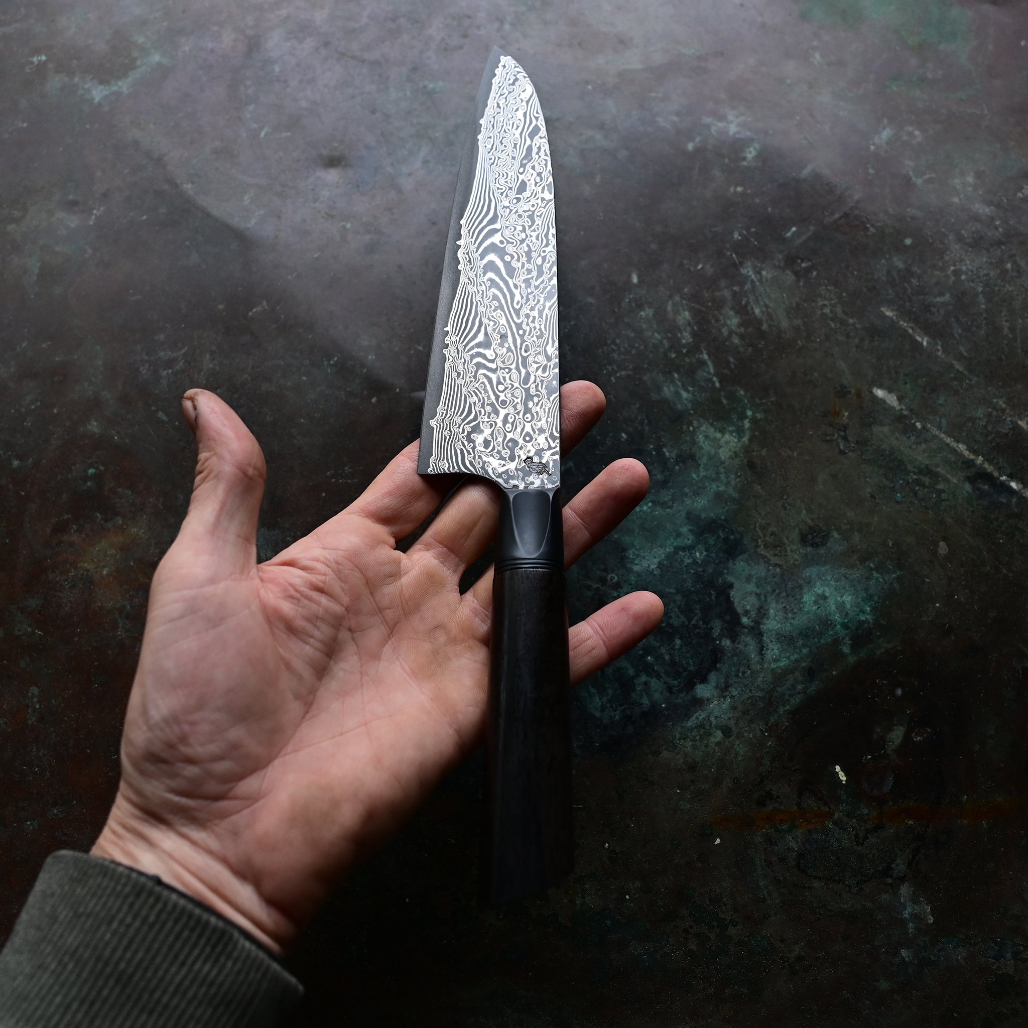 Hand-forged Santoku knife resting on a concrete background, featuring a deep black 7,000-year-old Irish bog oak handle, a contrasting silver anodized aluminum bolster, and a 7.5" VG10 stainless Damascus blade with intricate patterns.