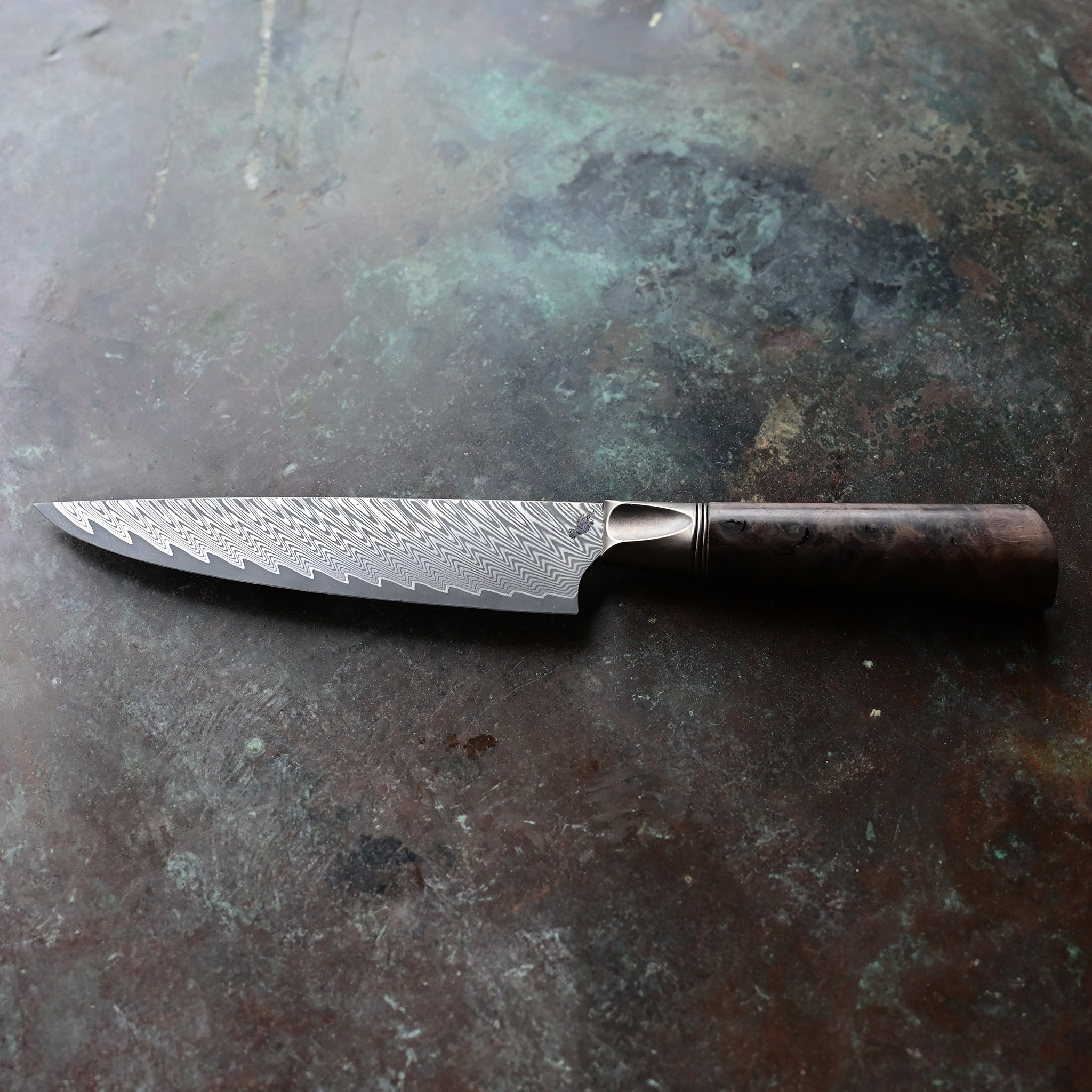 Distinctive 6.5" cutting edge petty knife on a concrete surface, showcasing a Zephyr Zigzag Damascus pattern, an aged bronze bolster, and a Claro walnut burl handle with rich brown and tan patterns