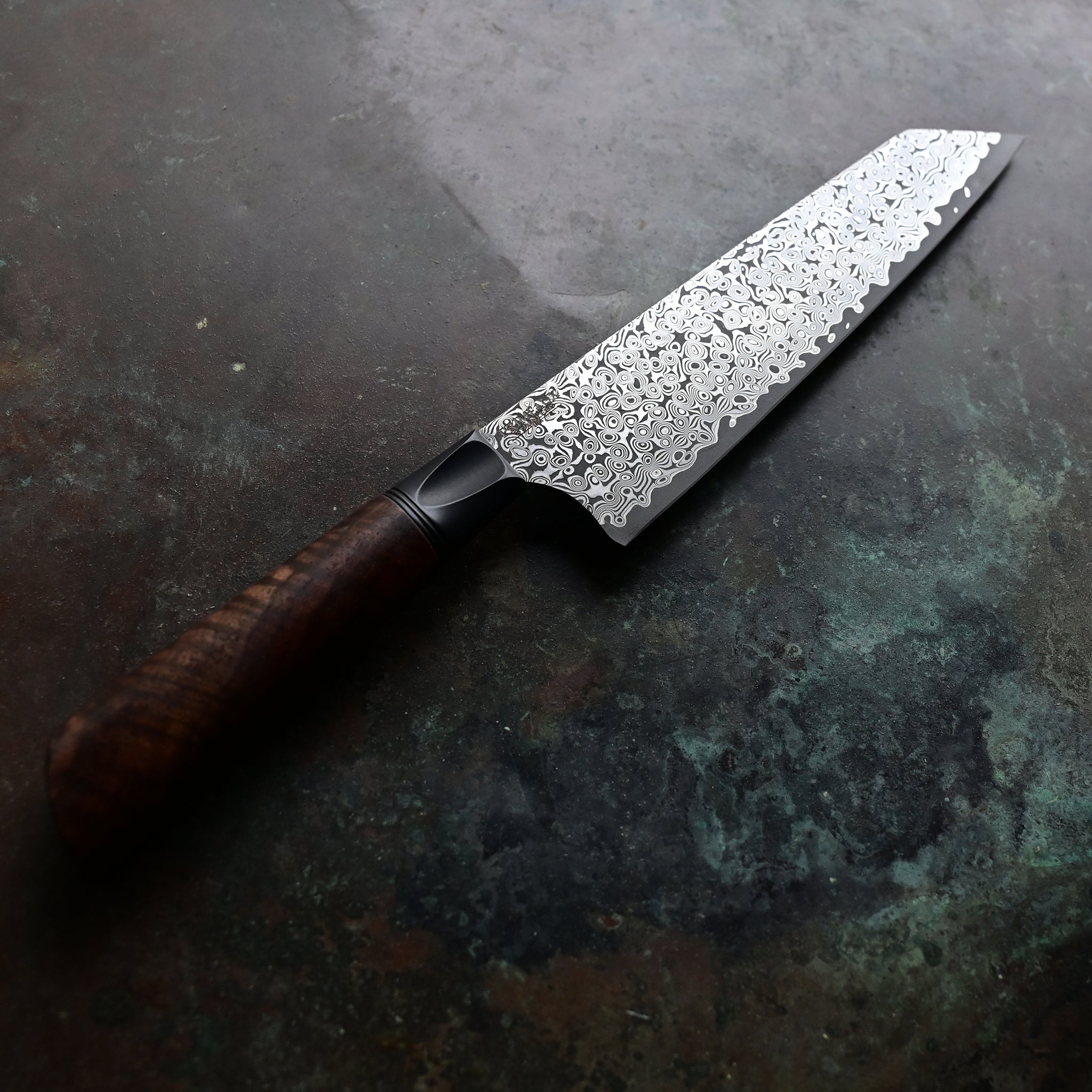 Elegantly redesigned Kiritsuke knife on a concrete surface, with a lighter, sharper blade, a warm brown Claro walnut handle, a sleek black anodized bolster, and a 7" Raindrop San Mai Damascus blade showcasing a mesmerizing pattern