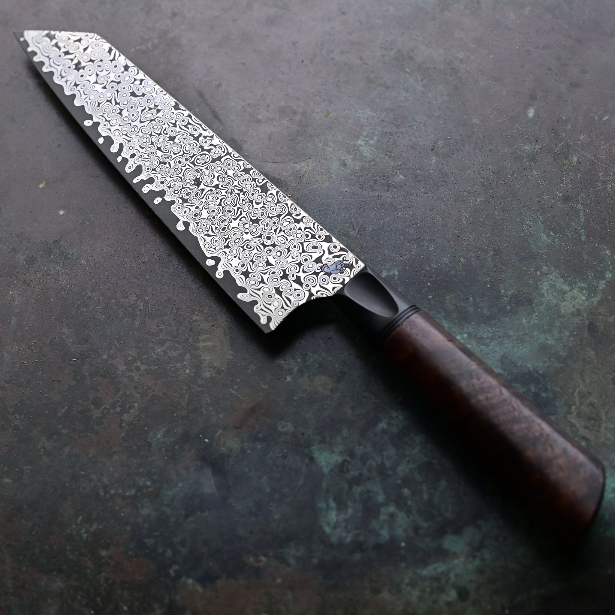 Elegantly redesigned Kiritsuke knife on a concrete surface, with a lighter, sharper blade, a warm brown Claro walnut handle, a sleek black anodized bolster, and a 7" Raindrop San Mai Damascus blade showcasing a mesmerizing pattern